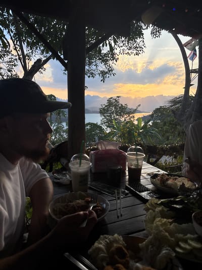 The image depicts an outdoor dining scene in Ban Ta Khun, Thailand. The setting appears to be a restaurant or cafe with a view of a lake or bay surrounded by trees and mountains. The sky is visible through the trees, showing a sunset with yellow and orange hues.
On the wooden table, there are several dishes of food, drinks in clear plastic cups with straws, and utensils. Two people are visible: one wearing a white shirt and a baseball cap, and the other person is partially visible. One person is eating with a fork.
A Thailand flag is visible at the top of the frame.