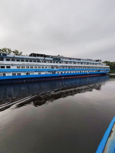 This is a full-shot, eye-level, outdoor, vertical photograph of a multi-deck passenger ship on a body of water. The ship is predominantly white with a blue stripe along its hull. It has numerous windows and balconies. The ship's name, "THE LITTLE PRINCE," is visible on its upper deck. A few individuals are visible on the upper decks. The water in front of the ship is dark and reflects the ship, with some ripples. Trees are visible on the far bank of the water. The sky is overcast. The location is identified as Vasil'ki, Russia.
