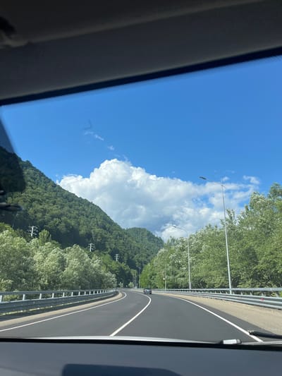 This image depicts a view from the interior of a vehicle looking out at a road curving through a mountainous, forested area. The sky is bright blue with scattered white cumulus clouds. On the left side of the road, a steep, densely forested mountain rises. On the right side, there is a line of trees and then more forested mountains in the distance. The road itself is asphalt, with clear white lane markings and a metal guardrail along the side. There are streetlights visible along the road, and power lines and poles can be seen on the mountainside. A few cars are visible on the road in the distance. The lower portion of the image shows the dashboard and windshield frame of the vehicle from which the photo was taken. The scene is likely a highway or major road in a rural, mountainous region. The location is identified as Kepsha, Russia.