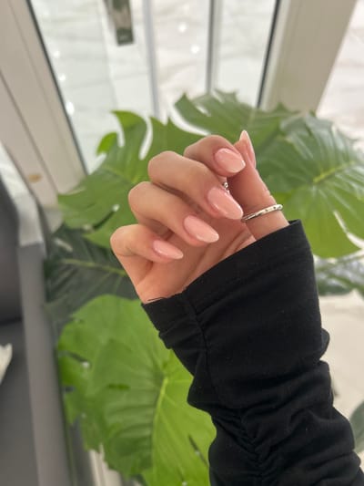 A hand with manicured nails, painted a pale pink color, is positioned in front of a leafy green plant. A simple silver ring adorns the ring finger. The hand is wearing a black, gathered-sleeve garment. The background includes a window and a glimpse of a blurred interior, suggesting an indoor location.