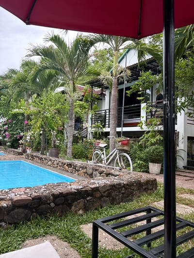 A tranquil outdoor scene unfolds at a resort in Bố Trạch Commune, Vietnam. In the foreground, a vibrant turquoise swimming pool is bordered by a rustic stone wall, with lush green grass growing around its edges. To the left, a red umbrella casts a shadow, hinting at a shaded seating area. A white bicycle with a basket filled with red flowers stands near the pool, adding a touch of charm. Behind the pool, a two-story building with white walls and balconies is surrounded by tropical foliage, including tall palm trees and flowering vines in pink and white. The sky is overcast, suggesting a warm, humid day.