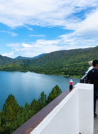 The image captures a scenic view of a large, tranquil blue lake surrounded by lush green, undulating hills under a partly cloudy sky. In the foreground, a white railing with a dark wooden top is visible, serving as a vantage point. A single bottle of Coca-Cola rests on the wooden railing. Two individuals are present, their backs partially turned to the camera. One person, wearing a black t-shirt with a graphic on the back, stands near the railing, looking towards the lake. To their left, another person, wearing a light-colored hijab and a white top, is also observing the landscape. The environment suggests a natural setting, possibly a viewpoint or resort overlooking the lake. The location is identified as Turpuk Sihotang, Indonesia, a region known for its natural beauty.