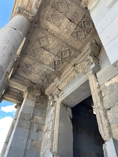 The image shows a close-up, low-angle view of the entrance to the Temple of Garni in Garni, Armenia. The temple, constructed of grey basalt blocks, features intricate stone carvings on its facade and ceiling. The ceiling is adorned with geometric patterns and floral motifs carved into individual stone panels. A large, fluted column stands to the left of the entrance, partially obscuring the sky, which is blue with scattered white clouds. The entrance itself is a large, arched doorway framed by carved stone. The weathered stone suggests the age of the structure. The light suggests it is daytime.
