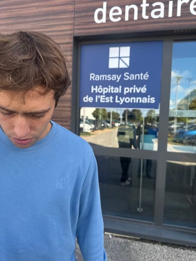 An outdoor image captures a young male in the foreground, positioned slightly to the left, looking downwards with his head tilted. He has light brown, wavy hair and wears a plain light blue crew-neck sweatshirt featuring an embossed "CH HC" logo on the upper left chest. Behind him is the glass entrance of a building, identified as a medical facility in Saint-Priest, France. Above the doors, a partial sign reads "dentaire." A prominent blue panel on the glass doors displays a white square logo and the text "Ramsay Santé / Hôpital privé / de l'Est Lyonnais." The reflective glass reveals a parking lot scene beyond, containing several parked vehicles and indistinct reflections of at least two individuals. The building's exterior around the entrance features horizontal brown paneling. The overall lighting suggests daylight conditions.