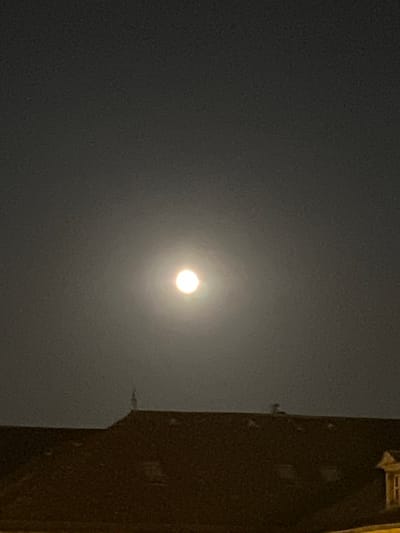 A full moon is visible in a dark, hazy sky above the rooftops of Espalion, France. The moon, appearing as a bright, glowing orb with a visible halo, is positioned slightly left of the center of the frame. The foreground features the dark silhouette of a building's roof, with visible dormer windows and architectural details. The overall scene conveys a nighttime ambiance in an urban setting.