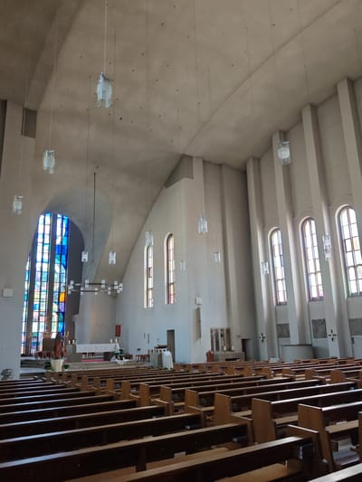 This is a photograph of the interior of a modern church, likely located in Memmingen, Germany. The church features a high, vaulted ceiling with numerous minimalist, cylindrical pendant lights. The walls are a pale, concrete-like material, with tall, arched windows filled with stained glass, primarily in shades of blue, red, and yellow, visible on the left side. To the right, a series of tall, narrow windows with divided panes allow natural light to enter. The seating area consists of rows of dark wooden pews arranged in a nave facing an altar area. The altar itself is elevated and covered with a white cloth, flanked by candles and a statue. The overall aesthetic is one of spaciousness and minimalist design, typical of mid-to-late 20th-century religious architecture.