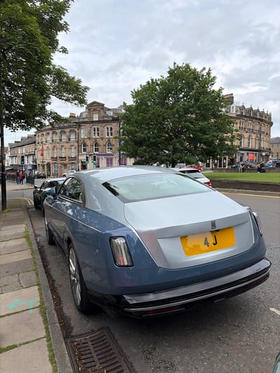 An outdoor, slightly rotated vertical shot captures the rear-end of a luxurious, two-tone Rolls-Royce car parked curbside on a cloudy day in Harrogate, United Kingdom. The car is primarily dark blue or black at the bottom and a lighter grey or silver on the top half, with a prominent yellow UK license plate partially visible, showing "4 J". The Rolls-Royce emblem is clearly visible on the trunk.

The car is parked on a paved street next to a concrete sidewalk. To the left of the car, a lush green tree with dense foliage occupies a significant portion of the mid-ground. Beyond the tree and the street, a line of stately, light-colored stone buildings with ornate architectural details, such as arched windows and decorative cornices, stretches into the background. Some of these buildings appear to house businesses, with a "NatWest" bank sign visible on one, and a "Costa Coffee" sign partially visible further down the street.

In the distance, several people are seen walking on the sidewalks or gathered on a grassy area, suggesting a typical daytime urban scene. Traffic lights are visible further down the street, one showing a green signal. The sky is overcast with thick grey clouds, indicating a cloudy or possibly gloomy day. No specific activity beyond cars being parked and people walking is evident. The overall impression is that of a quiet street scene in a historic British town, characterized by elegant architecture and modern luxury.