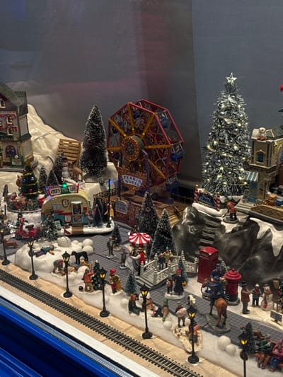 This media file, captured in Clamart, France, presents a vibrant and intricate miniature Christmas village display, illuminated as if at night. The scene is set on artificial snow-covered terrain, depicting a festive winter market or fair bustling with activity.
In the upper central area, a prominent, silver-flocked miniature Christmas tree stands tall, adorned with numerous bright lights and topped with a star. To its left, a colorful miniature Ferris wheel dominates the mid-ground, featuring gondolas and a central hub that are also lit. The Ferris wheel structure clearly displays the text "FERRIS WHEEL" in white lettering.
Numerous small figurines populate the village, depicting people of all ages, dressed in winter attire, engaged in various activities like walking, gathering, and enjoying the festive atmosphere. A figurine resembling Santa Claus is visible near the bottom center. Animal figurines include horses pulling a carriage in the upper right quadrant and several reindeer-like figures.
Pathways within the village are lined with miniature lampposts, casting a warm glow. Various small buildings and stands are scattered throughout the scene. One notable structure is a camper-like vehicle with a sign that reads "TED'S TREE LOT," indicating a place for selling Christmas trees. Another sign, displaying "Merry Christmas," is visible near a decorated tree in the upper right. Additional details include a red fire hydrant and a red mailbox.
The right side of the image features miniature train tracks, suggesting a functioning train display that enhances the dynamic nature of the village. The overall impression is one of a cheerful and bustling holiday celebration, brought to life through intricate details and warm, inviting lighting, creating a magical winter wonderland scene.