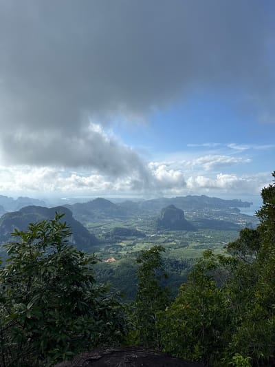 The image captures a panoramic vista from a high vantage point, overlooking a lush, mountainous landscape and a coastal town. The foreground is dominated by the dense foliage of green trees and the rough texture of dark rock, suggesting the viewpoint is from a cliff or a high peak. In the mid-ground, rolling hills covered in verdant vegetation stretch out, interspersed with patches of cultivated land and scattered buildings that form a small town. Further in the distance, dramatic limestone karsts rise from the landscape, some with sheer cliffs and others with more rounded, vegetated slopes. The horizon is marked by a body of water, likely the Andaman Sea, with several islands visible in the distance. The sky is a mixture of dramatic, heavy grey clouds and patches of bright blue sky with scattered white clouds, indicating dynamic weather conditions. The scene is evocative of Krabi province in Thailand, known for its dramatic karst topography and beautiful coastal scenery, with the town possibly being Ao Nang or a similar settlement in the Ban Din Daeng Nui area. No people are visible in the image.