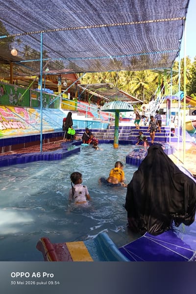 The image depicts a group of people enjoying a pool at what appears to be a water park. The location is likely in Palohseulimeng, Indonesia. Several children are in the pool, some are swimming, and others are standing or wading in the water. There are adults present, some of whom are wearing hijabs. In the background, there are colorful slides, stairs, and other water park features. A mesh-like structure provides shade over the pool area. The photograph was taken with an OPPO A6 Pro on May 30, 2026, at 09:54 AM.