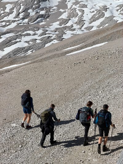 The image depicts four individuals hiking uphill on a scree slope. The terrain is predominantly covered with small stones and gravel, with patches of snow and rock formations visible on the steeper incline in the background. The individuals are equipped with backpacks and trekking poles, suggesting an outdoor excursion. They are walking away from the camera, towards the upper part of the slope. The lighting indicates it is daytime, with shadows cast by the hikers and the terrain. The environment appears to be mountainous, likely in the Austrian Alps, potentially in or near Wildermieming, Austria.