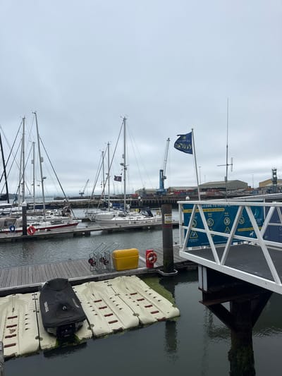The image shows a marina with several sailboats docked. In the foreground, there are floating platforms with a covered personal watercraft. A yellow storage bin and a life preserver are visible on a wooden dock. A white ramp leads to a blue sign that reads "WELCOME TO POOLE QUAY" and lists various regulations, including "NO SWIMMING" and "NO FISHING." A blue flag with a logo is flying from a flagpole. In the background, cranes and industrial buildings suggest a port or harbor area. The sky is overcast, and the water is calm. The scene appears to be the Poole Quay in Poole, United Kingdom, a popular area for boating and recreation. There are no people actively engaged in any activity in the image.