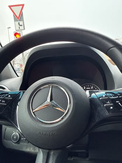 The image provides a first-person view from inside a Mercedes-Benz vehicle, looking through the windshield. The foreground is dominated by the black leather steering wheel, centered with a chrome Mercedes-Benz star logo and the embossed word "AIRBAG." Controls are visible on both spokes of the steering wheel.
Outside the vehicle, under an overcast sky, an intersection or street environment in Hildesheim, Germany, is depicted. A red traffic signal is illuminated directly ahead, indicating a stop. Above it, a red-bordered white triangular "Give Way" sign is present, with a white rectangular sign depicting a black left-turn arrow mounted beneath it. In the mid-ground, a commercial building is visible, identifiable as a "Suzuki" dealership, further pinpointing the location to a commercial automotive district within Hildesheim, such as the vicinity of Cheruskerring 24 (Autohaus Südhannover GmbH). Bare trees are visible in the background, consistent with winter or early spring. No individuals are explicitly visible.