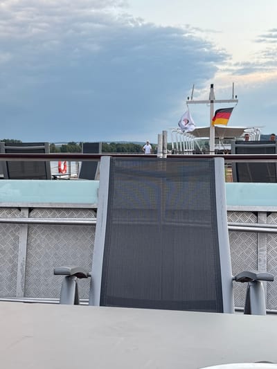 The image shows a scene from what appears to be a river cruise ship deck. In the foreground, a gray mesh lounge chair is partially visible, with a metallic support arm attached to a textured metal floor. Behind the chair, more lounge chairs and railings are visible, suggesting an outdoor seating area.
Further back, a German flag and another white flag with a logo are flying from a mast on the ship. A few people are visible on the deck, one standing near the flags and another in the distance. A life preserver is attached to a railing. The background shows a lush green treeline against a cloudy sky, indicating an overcast day. The overall scene suggests a leisurely moment on a boat, likely during the daytime. The location context provided is Winkel, Germany.