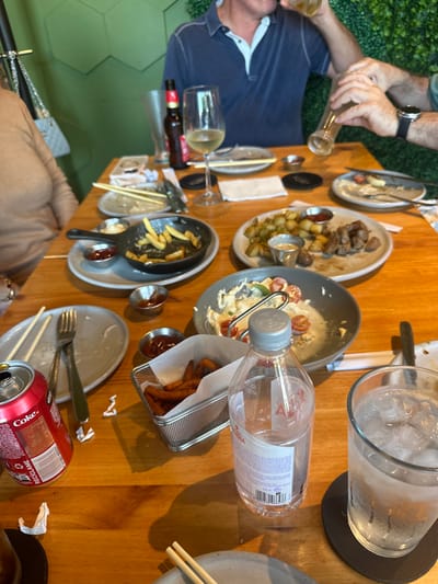 A wooden table is covered with remnants of a meal. Dirty plates, some with forks or chopsticks, scatter the surface. There are also metal containers holding sauces and other foods. Bottles of water and soda, along with a glass of ice water, are visible. Two people sit partially at the table, one with a blue shirt and the other with a beige jacket. A beer bottle and a wine glass are also placed on the table.