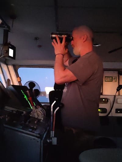 A man with a gray beard and short, receding gray hair stands inside a ship's control room. He is holding a cylindrical object to his eyes, possibly binoculars, and looking out of a large window. The room is dimly lit, with various control panels and instruments visible. A small fan is positioned near the window. An exit sign is illuminated on the right side of the frame. The man is wearing a gray t-shirt and a thin bracelet on his left wrist. The location is unknown, and there's no verifiable indication of Kostyaevka, Russia.