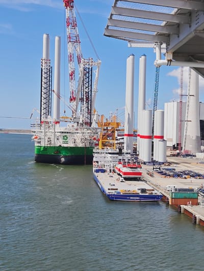 The image depicts an industrial harbor scene under a clear blue sky, suggesting a sunny day. In the foreground, a large body of water is visible, with moderate ripples indicating a light breeze. Moored to the right is a blue barge carrying several large containers, one green and one orange, secured by ropes. The barge has the text "BRAVEWIND" in white lettering along its side.
Dominating the mid-ground is a massive green and white vessel, a specialized offshore construction ship, with several tall, white cylindrical towers (likely components for wind turbines) loaded onto its deck and being supported by extensive scaffolding and cranes. A large red and white crane is prominently featured, extending high into the sky. Behind this vessel, more of these cylindrical towers are visible, some standing vertically and others lying horizontally, along with various other industrial structures and equipment, including more cranes of yellow and blue colors. In the background, a distant, flat coastline can be seen under the expansive sky. The scene appears to be a bustling port or shipyard involved in the construction or transport of large industrial components, likely related to offshore wind energy given the presence of the wind turbine towers and the context of Esbjerg, Denmark, a major hub for the offshore wind industry. There are no visible people in the main focus of the image.