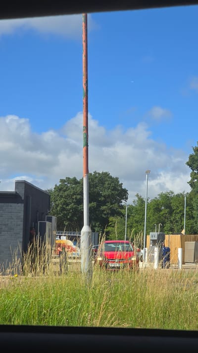 This is an eye-level, medium-distance outdoor shot of a red car at a gas station in Wicken, United Kingdom. The car is positioned in front of a pump, with a wooden fence and another structure behind it. Tall grass is in the foreground, obscuring the bottom portion of the scene. A tall, weathered pole, painted red and green, stands to the left of the car. A building with dark grey brickwork is visible to the left of the pole. The sky is blue with scattered white clouds, and a faint airplane trail is visible in the upper right portion of the sky. The overall lighting suggests it is daytime.