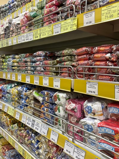 The image shows a shelf in a supermarket filled with an assortment of wrapped candies. The shelves are metal, and the candies are arranged in plastic packaging of various colors and designs. Many of the packages are transparent, revealing the candy inside. Some shelves have yellow price tags with Cyrillic text and numbers, indicating prices and possibly product names. The background is out of focus but suggests more shelves and products in the store. The overall impression is one of a well-stocked candy aisle.
