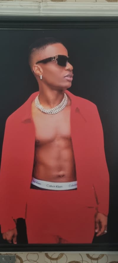 The image displays a head-to-mid-torso shot of a man, likely a celebrity, posing against a plain black background. The photograph, which appears to be a framed print, is currently rotated 90 degrees counter-clockwise.
The man, who has dark skin and short, neatly cut hair, is wearing dark sunglasses that obscure his eyes. He has a slightly open mouth, as if about to speak or in a relaxed pose. A prominent, multi-layered silver or white chain necklace adorns his neck. He is dressed in a vibrant red blazer or jacket, which is open, revealing his bare chest and abdomen. Below his navel, the waistband of a pair of boxer briefs is visible, clearly displaying the brand name "Calvin Klein" in white text on a dark band. His left hand, adorned with a ring on one finger, is slightly visible towards the right side of the frame.
The overall scene suggests a professional photoshoot or promotional image, emphasizing a stylish and confident demeanor. The lighting appears to be controlled studio lighting, highlighting the subject against the dark background. The context provided indicates this image is located in Alasia, Nigeria, although no specific environmental cues within the image itself confirm this.