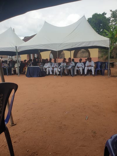 The image depicts an outdoor event, likely in Nkwerre, Nigeria. A large white tent provides shade for rows of seated individuals. The attendees appear to be dressed formally; some wear white garments with red accents, while others don various outfits. A table covered with a blue cloth is visible in the center. Blue plastic chairs accommodate the guests. The ground is reddish-brown soil. A building with arched openings is situated behind the seated individuals. Foliage and trees are visible in the background.