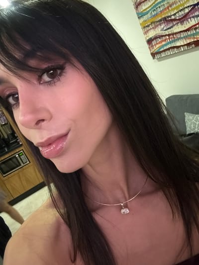 A woman with dark hair and fringe gazes into the camera. She is wearing a fine gold necklace with a single diamond pendant. Her skin is fair and lips glossy. The background is softly blurred with hints of a colorful abstract artwork on the wall and a glimpse of kitchen appliances.