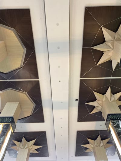 The image shows a low-angle shot of a modern ceiling with intricate geometric designs. The ceiling is composed of dark wood panels with starburst patterns made of light-colored wood and silver trim. In the lower left and right corners, marble-clad columns with decorative green tile accents are visible. The overall aesthetic suggests a public space, possibly a hotel lobby or a commercial building, in Aluekambuk, Indonesia. The lighting is bright and appears to be from recessed ceiling lights. There are no people visible in the image. The scene is clean and well-maintained, with a focus on architectural detail and design.