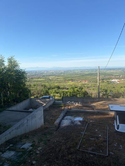 A landscape photograph taken from an elevated vantage point, likely in or near the city of Brestnik, Bulgaria, depicts a panoramic view spanning from a clear blue sky to a distant cityscape nestled amidst lush green vegetation. A utility pole with overhead wires stands prominently in the middle ground, visually connecting the foreground elements with the sprawling landscape below. The foreground features a concrete structure or retaining wall along with overgrown vegetation, suggesting an abandoned or undeveloped area. A metal frame or enclosure lies on the ground amidst dried grass, partially obscured by debris. A small white plastic chair sits near a flat white surface, possibly a section of a building or a large slab of concrete. The composition suggests a scene of observation or overview, capturing a blend of urban development and natural environment.