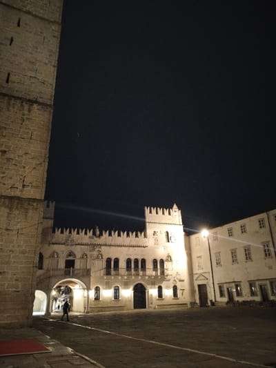An illuminated building with intricate carvings and arched windows stands against a dark sky. A taller, blocky stone structure is partially visible on the left. A person walks through an archway beneath the building. A light shines brightly near the building. The foreground is a paved square with lines running through it, and a red mat. Other buildings are visible to the right. The scene is dimly lit.