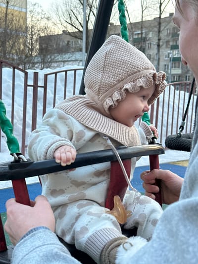 The image depicts an infant seated in a red and black baby swing at an outdoor playground in Sadovaya, Russia. The infant, positioned centrally, wears a light beige knitted bonnet with a ruffled brim, a matching ribbed collar, and a cream-colored knitted romper adorned with light brown heart patterns. Light-colored, possibly fur-lined, booties cover the feet. A pacifier, attached by a spiral cord and white clip, hangs from the infant's romper near the left leg. The infant's left profile is visible, showing fair skin and an upward-curved expression, directed towards an unseen individual. The hands of an adult, wearing a light grey long-sleeved garment, are visible on the right, gripping the red support bars of the swing, indicating interaction with the infant. The ground is covered with white snow, with sections of a blue and light-colored padded surface beneath the swing. A dark brown metal fence is partially visible behind the swing, also partially covered by snow. In the background, leafless deciduous trees and multi-story residential buildings are discernible under a pale sky.