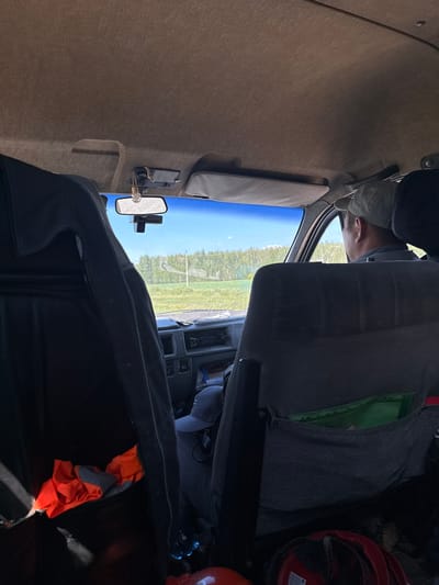 The image appears to be taken from inside a vehicle, likely a truck or van, looking towards the front windshield and passenger side. The interior of the vehicle is visible, with tan-colored upholstery on the ceiling and parts of the dashboard. A person, partially visible, is wearing a cap and appears to be looking out the windshield.
Outside the vehicle, a clear blue sky suggests fair weather. A landscape of green fields and trees is visible through the windshield, indicating a rural or semi-rural setting. A utility pole can be seen in the distance, a common feature in such areas.
Inside the vehicle, a rearview mirror is mounted on the windshield. There is also a black dashboard with some visible buttons or controls. On the passenger side, a dark gray seat is prominent, with a bright green material peeking out from a pocket or compartment. Towards the right side of the image, a red safety helmet is visible, suggesting the occupant might be involved in work or an activity requiring protective gear.
Further back in the vehicle, a large black bag or cover is present, with a bright orange garment folded on top of it. The lighting suggests it is daytime, with natural light coming from outside.
The overall impression is a snapshot from the interior of a vehicle during a daytime drive through a rural landscape in Kazakhstan, possibly related to some form of fieldwork or construction activity. No visible text is discernible in the image.