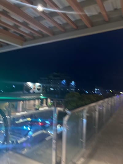 The image depicts a blurry night scene from a high vantage point, possibly an elevated walkway or bridge, overlooking a city street in Davao City, Philippines. The foreground shows a metal railing and a portion of a pathway. Below, streaks of light from vehicles indicate movement on the street, with prominent blue, red, and white lights suggesting traffic signals and headlights. In the background, illuminated buildings and trees are visible against a dark night sky. A large, out-of-focus billboard or signage is present on the left side of the scene. The overall impression is one of a dynamic urban environment at night.