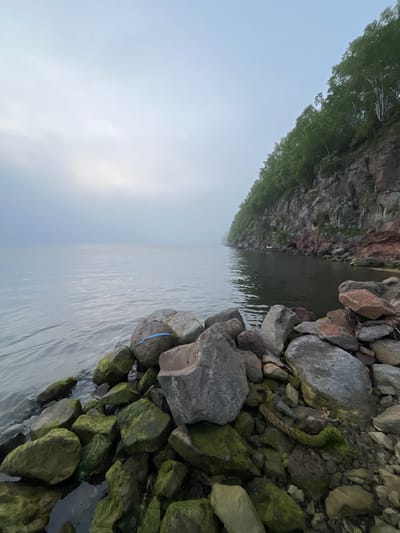 The image shows a rocky shoreline on a misty day. The water is calm and reflects the overcast sky. A steep, forested cliff rises on the right side of the frame. In the foreground, a pile of large rocks, some covered in green moss, are visible. A blue ribbon or strap is tied around one of the rocks. The overall scene suggests a natural, undeveloped coastal area.