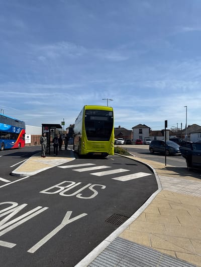 This image depicts a modern bus station or designated bus stop area in Salisbury, United Kingdom, under a clear blue sky with sparse clouds. The scene is dominated by two double-decker buses.
In the foreground, a bright lime green double-decker bus, bearing the registration "WK66 CYD" and "STONEHENGE Buy tickets online" text on its rear, is parked facing away from the viewer at a bus stop. To its left, partially obscured, is a red and blue double-decker bus with "X3" visible on its side. Between the two buses, a dark-colored bus shelter is present, with several individuals standing near it, seemingly waiting for or boarding the green bus. One person in a grey tracksuit with red-soled shoes stands clearly visible on the paved island adjacent to the bus shelter.
The road surface is fresh black asphalt, marked with white pedestrian crossing stripes leading to a raised central island. The words "BUS ONLY" are painted in large white letters on the asphalt, with "ONLY" partially visible in the bottom left. The central island and surrounding pedestrian areas are paved with light-colored slabs, incorporating grey tactile paving at the curb edges. A square metal drain grate is visible in the asphalt near the curb.
In the background, beyond the bus stops, residential or commercial buildings with pitched roofs are visible, along with multiple parked vehicles, including a dark blue SUV and a white SUV. Several streetlights are visible throughout the scene. The overall impression is a functional, recently developed urban transportation hub.