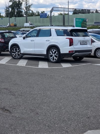 This media file shows a parking lot with several cars parked in it, on a sunny day. The main subject is a white SUV parked in the foreground, with its rear and side visible. The SUV has a license plate that reads "K433AM 977". To the left of the SUV, there is a green sign with text in Russian. The sign appears to be an environmental awareness message, stating "Бережно к людям, бережно к природе" which translates to "Careful with people, careful with nature". Further to the left, there are more signs, one of which indicates directions to "МОСКВА" (Moscow) and "ЛОБНЯ". In the background, there is a green sound barrier fence, trees, and a cloudy sky. The scene appears to be in a suburban or highway rest stop area, given the presence of traffic signs and the sound barrier. The overall impression is a mundane scene of vehicles parked on a bright day, with hints of location information and a public service message. The image is oriented vertically, with the cars appearing sideways.