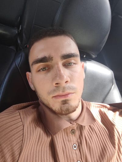 The image is a close-up selfie of a young man with light brown hair, prominent eyebrows, and green eyes. He has a light beard and a few freckles on his nose. He is wearing a ribbed, collared, button-up shirt in a light brown or tan color. The background appears to be the interior of a car, with dark leather seats visible. Sunlight is illuminating the scene from the right side, creating highlights on the man's face and the car seat. The image does not provide any information about the location in the city of Khorol, Russia.