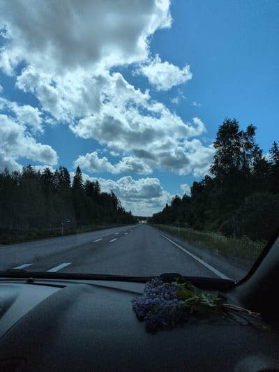 The image depicts a daytime scene from the interior of a vehicle traveling on an asphalt road in Heinola, Finland. The perspective is forward-facing through the windshield, with the dark dashboard occupying the lower portion of the frame. A small bunch of purple flowers, consistent with lilacs, rests on the passenger side of the dashboard.

The road, marked with white dashed lane lines and a solid white shoulder line on the right, extends into the distance, appearing to curve slightly. Dense, dark green forest, comprising both coniferous and deciduous trees, lines both sides of the roadway, forming a continuous natural barrier. White road marker posts are visible along the right shoulder.

The sky above is partially clear, exhibiting a bright blue hue, and partially covered by scattered white cumulus clouds of varying sizes. Darker, heavier clouds are present higher in the sky. Natural light illuminates the outdoor scene, while the car's interior, specifically the dashboard, appears comparatively darker. No individuals are visible in the image.