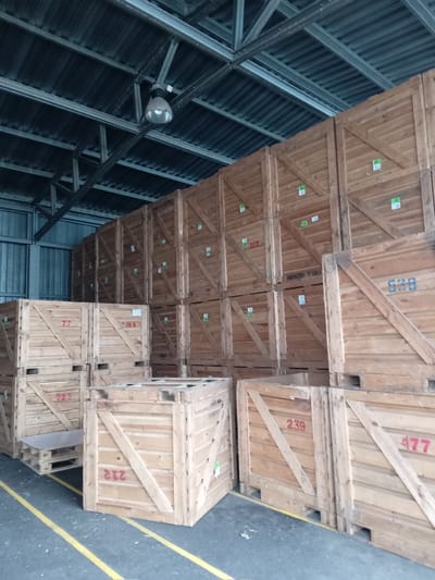 The image shows a storage facility in Biloziria, Ukraine, filled with large wooden crates stacked high. The crates are of various sizes and appear to be used for storage or transport. Some crates have numbers or labels painted on them, such as "77", "446", "525", "212", "239", "477", and "538". The facility has a metal roof with exposed beams and industrial lighting fixtures. The floor is concrete with yellow lines painted on it, possibly marking pathways or storage areas. The overall impression is one of a well-organized, functional warehouse space.