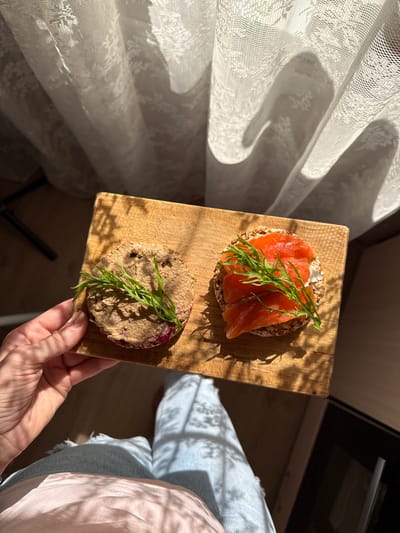 A top-down, medium shot captures a person's hand holding a wooden cutting board with two open-faced sandwiches. The sandwiches are on round, grainy crackers. One sandwich is topped with a beige spread and garnished with green herbs. The other sandwich features sliced red fish, a white spread, and green herbs. Sunlight streams in from the right, casting shadows of the curtain and foliage onto the scene. The background shows a white, lace-patterned curtain. The person is wearing light blue jeans with ripped knees and a pink shirt. The setting appears to be indoors, possibly a kitchen, in Kavkazkaya, Russia.