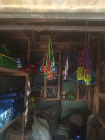 The image displays the interior of a retail or storage space within a building in Kofar Ruwa, Nigeria. The scene is characterized by dim illumination and a dense arrangement of household cleaning supplies. Prominently suspended from an overhead beam are multiple clusters of plastic brushes. The left cluster features brushes with green handles and multi-colored bristles (red, blue, yellow), while the right cluster comprises brushes with blue handles and green, yellow, and red bristles. A smaller grouping of brushes with white and black handles hangs centrally.
Along the left side, wooden shelving units are visible. The top shelf holds stacked green and blue plastic items, consistent with basins or buckets. A lower shelf contains numerous stacked blue plastic goods, some appearing individually wrapped. Several silver-colored poles, likely handles for cleaning tools, lean vertically against these shelves. In the background, additional wooden shelves house more merchandise, though specific items are less discernible. At floor level on the left, a large, light-colored sack is present, while other bundled items and green plastic containers are visible on the right. No individuals, actions, or direct interactions are depicted within the frame.