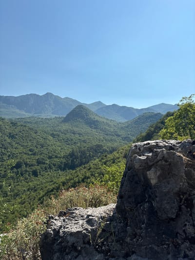 The image depicts a mountainous landscape under a clear blue sky. In the foreground, a large, textured gray rock formation dominates the right side of the frame, with dry, sparse vegetation growing around its base. Beyond the rock, a vast expanse of rolling, densely forested mountains stretches into the distance, their peaks fading into a hazy blue. The sunlight suggests it is daytime, likely morning or afternoon, with good visibility. The scene conveys a sense of natural grandeur and remoteness. There is no text or identifiable activity occurring. The location context points to Dupilo, Montenegro, a region known for its rugged terrain and natural beauty.