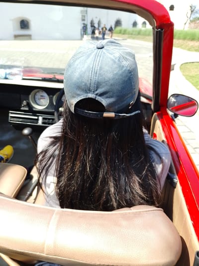 The image shows a person wearing a blue baseball cap and a grey shirt, viewed from behind, sitting in the passenger seat of a red convertible car. The person has long, dark hair. The car's interior includes a tan leather seat, a dashboard with gauges, and a gear shift. Outside the car, a paved area with a white building in the background is visible. The lighting suggests a sunny day. The location is identified as Bejalen, Indonesia, though this context is not visually evident in the provided image.