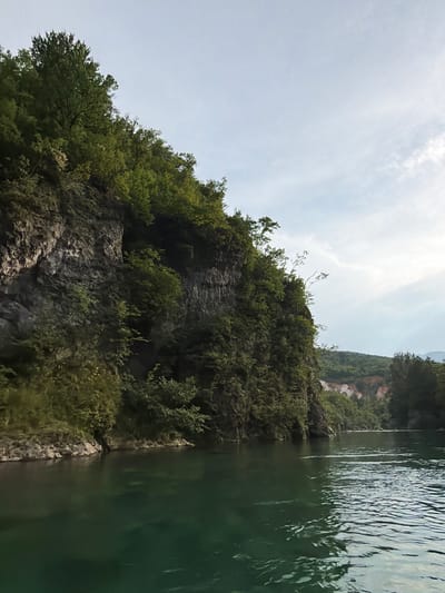 The image depicts a scenic view of a steep, rocky mountainside covered in lush green vegetation, which plunges down to a body of dark, rippling water. The water occupies the right side of the frame, its surface disturbed by ripples that catch the light. The mountainside, on the left, is densely forested with trees and smaller shrubs, their leaves a vibrant green. The rocks are rugged and appear to form a natural cliff face. The sky above is bright and overcast, with patches of lighter clouds and a hazy sun, suggesting it is daytime, possibly late morning or early afternoon.
The perspective suggests the photo was taken from a boat or a similar vantage point on the water, looking upwards towards the mountain. The overall impression is one of natural beauty and perhaps a sense of the wild or untouched landscape. There are no people or man-made objects visible, focusing entirely on the natural environment. The scene evokes a feeling of tranquility and the power of nature. No text is visible in the image. The location is Polje Bijela, Bosnia and Herzegovina, a region known for its natural landscapes and waterways.