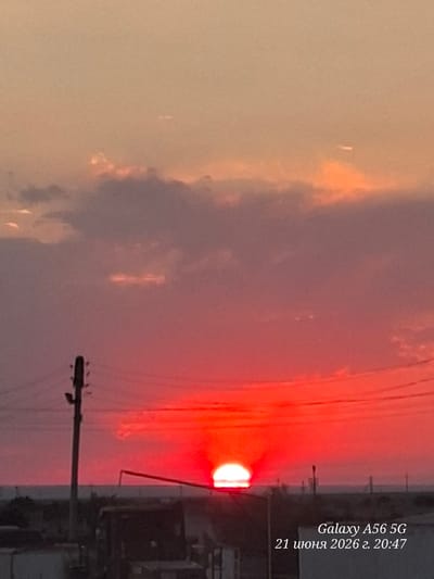 A wide, horizontal, full-shot image captures a vibrant sunset over a rural landscape. The sun, a brilliant orb of white-hot light, is partially obscured by the horizon, casting a fiery red glow across the sky. Wispy clouds, tinged with pink and orange, drift lazily above. In the foreground, silhouetted against the sunset, are several utility poles and power lines, their dark forms stark against the bright sky. In the distance, a low-lying structure and a vehicle are barely visible, suggesting a settlement or industrial area. The overall impression is one of peaceful, natural beauty, with a hint of human presence. There are no people visible in the image.