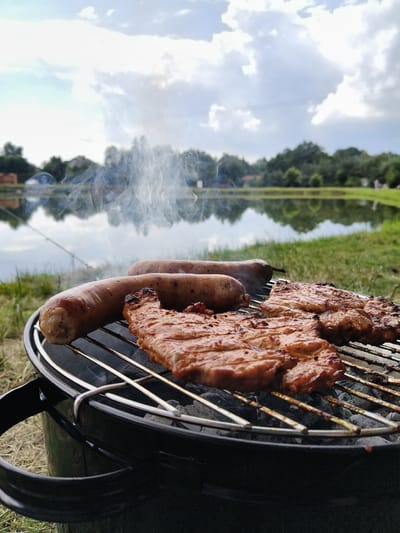 In the foreground, a black portable charcoal grill is visible, filled with gray coals. On the grill's grate, two sausages and several pieces of grilled meat, possibly pork steaks, are cooking, emitting wisps of white smoke. The grill is positioned on green grass. In the middle ground, a calm body of water, likely a pond or lake, reflects the sky and surrounding trees. A fishing rod is visible extending into the water. The background features a line of lush green trees and a cloudy sky with patches of blue. The scene appears to be set in a park or recreational area, consistent with the natural landscape of Ćwiklice, Poland. There are no people visible in the image.