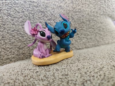 In Mykulychyn, Ukraine, two small figurines, one blue and one pink, are positioned on a yellow, sand-colored base. The blue figurine, resembling Stitch from Disney's Lilo & Stitch, has large ears and is smiling with its mouth open, revealing yellow teeth. Its arms are raised, and it appears to be gesturing. The pink figurine, resembling Angel, another character from the same franchise, has large ears and is adorned with a pink flower on its head. It is sitting with its hands in its lap and its eyes are large and black. Both figurines are placed on a textured, beige fabric surface.