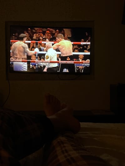 A person is lying in bed, positioned to the right of the frame, with their bare feet visible. Their legs are covered by dark pants and a flannel-patterned shirt. To the left, a television screen displays a boxing match with a crowd in the background. The boxing ring is indicated by ropes labeled "Scary Movie Boxing." The setting appears to be indoors, possibly a bedroom. There is no direct evidence within the image to confirm or refute a location in Genk, Belgium.
