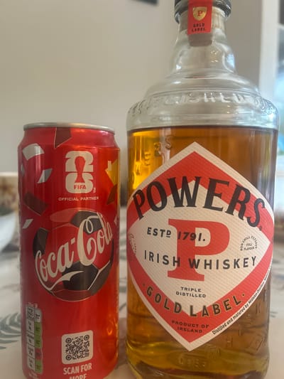 A close-up, slightly elevated shot captures a Coca-Cola can and a bottle of Powers Irish Whiskey. The Coca-Cola can, predominantly red, features a soccer ball graphic and "FIFA Official Partner" text. Nutritional information and a QR code are visible on the lower portion of the can. To its right, a clear glass bottle of Powers Irish Whiskey displays its iconic red and white label with "POWERS IRISH WHISKEY GOLD LABEL" prominent. The whiskey inside is a warm amber color. The bottle has a cork stopper and a red and gold foil seal. The background is blurred, suggesting an indoor setting, likely a table with a patterned tablecloth. The image does not provide any specific information about the city of Limerick or its location within it.