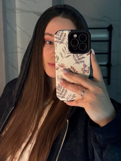 A female subject with long, dark brown hair and wearing a dark zippered hoodie is depicted taking a mirror selfie indoors. Her face is partially obscured by a smartphone held in her right hand, revealing her left eye, nose, and mouth. She exhibits light makeup, including pinkish eyeshadow. The smartphone, an iPhone with a triple camera array, is encased in a light-colored, vertically ribbed case adorned with botanical and butterfly patterns. Her right hand displays manicured nails and a silver-toned ring with a blue inlay on the ring finger. A light-colored, possibly floral-patterned top is visible beneath the hoodie. The background, reflected in the mirror, shows a light-colored wall, potentially tiled or paneled, with a dark, multi-bar towel rack mounted on the right side. The specific location within the city of Prikubanskii, Russia, is not discernible from the visual evidence.