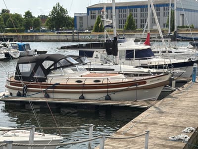 In Wismar, Germany, a fleet of sailboats and motor yachts are moored at a wooden dock on a sunny day. The foreground features a well-maintained motor yacht with a cream hull and dark brown trim, partially covered by a black canopy. The yacht is named "ALETTA DEUX" in gold lettering on its side. A wooden walkway, constructed from planks, extends into the water, with coiled ropes and mooring lines scattered on its surface. Behind this vessel, other boats are visible, including sailboats with tall masts and white hulls. In the background, industrial buildings with large windows and a grey facade line the waterfront, with a backdrop of lush green trees and a clear sky. The water is calm, reflecting the boats and the sky.
